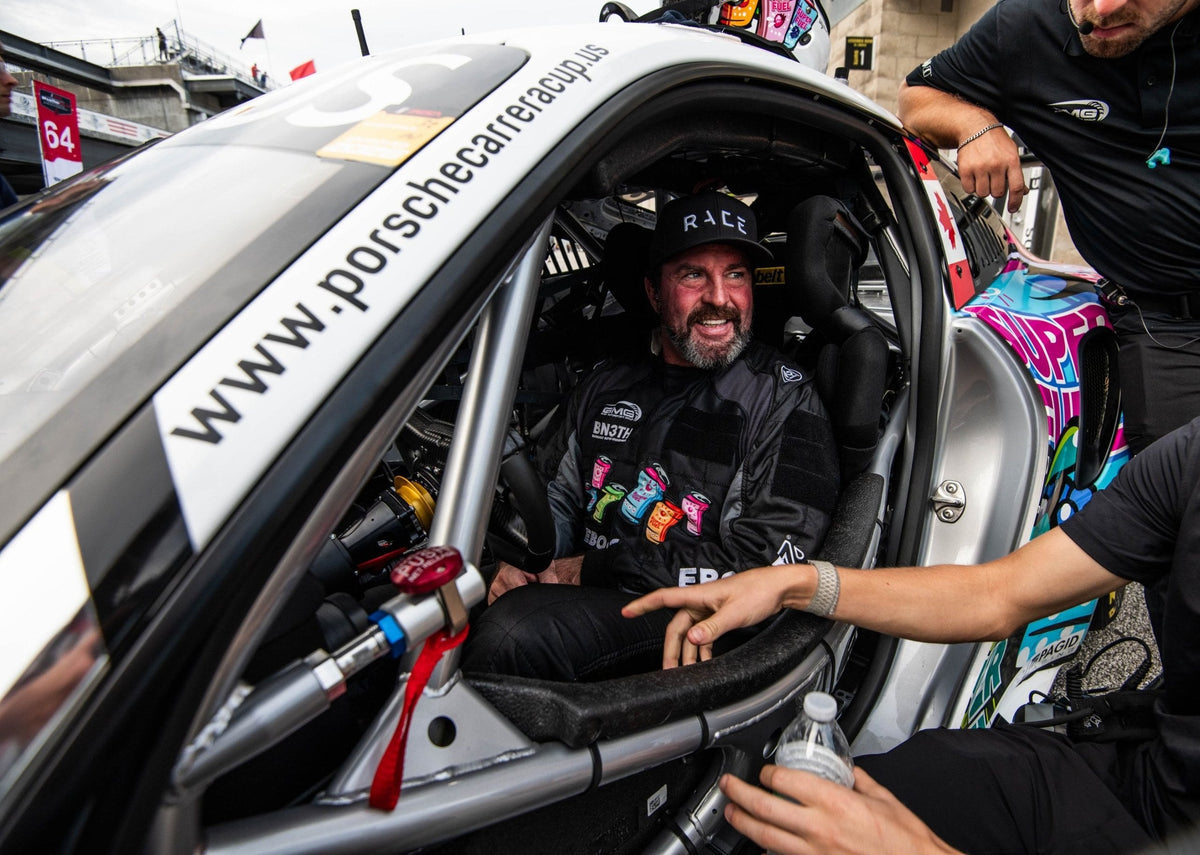 A driver in a GMG Racing suit seated in a Porsche Carrera Cup car as crew members lean in